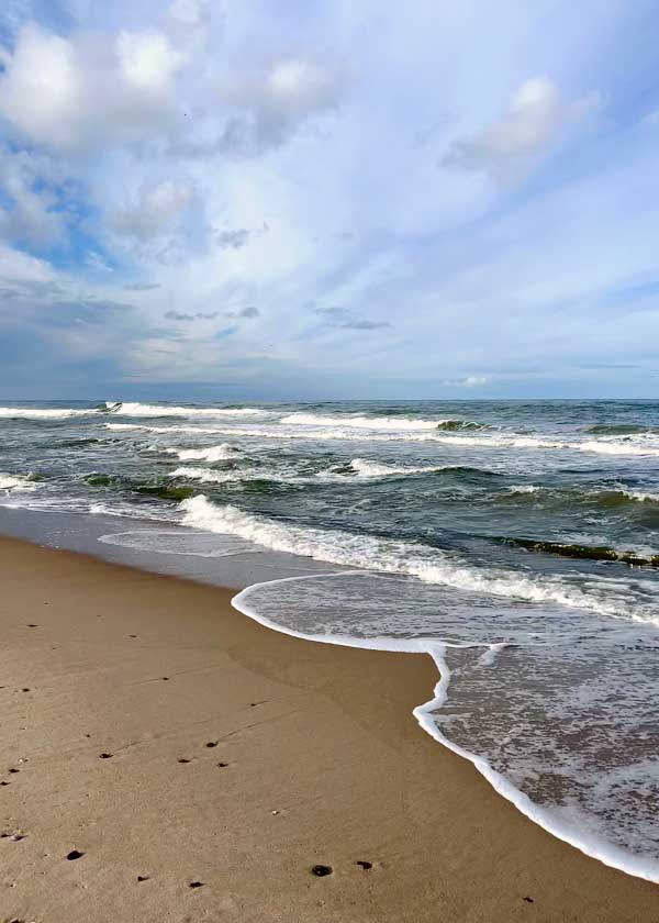 Sommerdag på stranden, bølgerne ruller sagte med strandkanten over dette himlen som viser en form hvor skyerne nærmest falder mod vandet